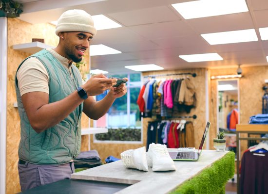 Male Sales Assistant In Pop Up Fashion Or Clothes Store Taking Picture Of Trainers On Mobile Phone